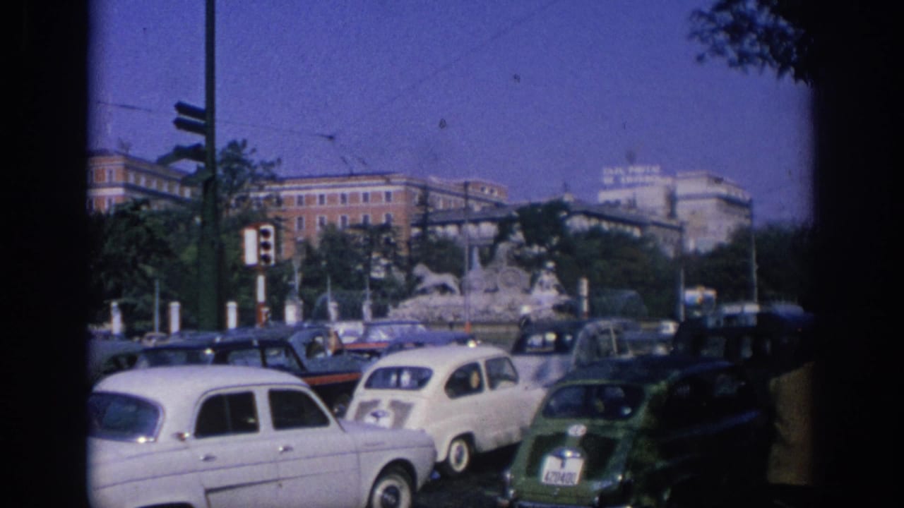 Busy street with compact cars, traffic lights, a fountain, and large buildings beyond.
