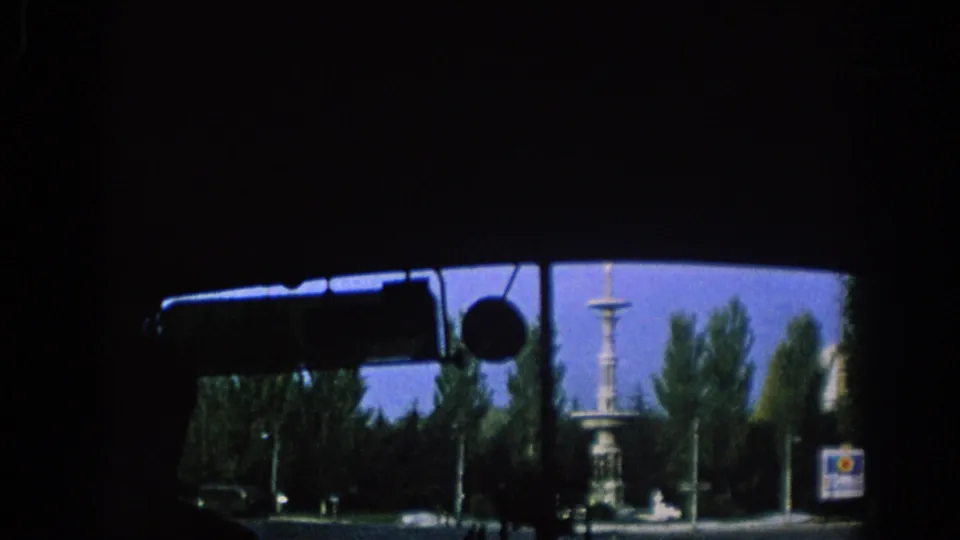 Passenger view from inside a moving car in Spain, 1966, with the windshield edge, rearview mirror, trees, street signs, and a distant monument visible.