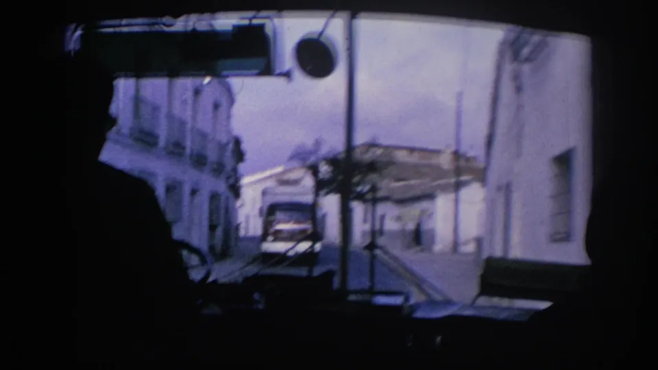 From inside a bus in Spain, dark seated figures frame a street where another bus appears ahead between white buildings.