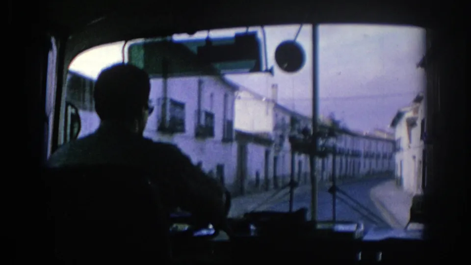 The driver is silhouetted from behind inside a bus, looking ahead at a narrow Spanish street with white buildings on both sides.