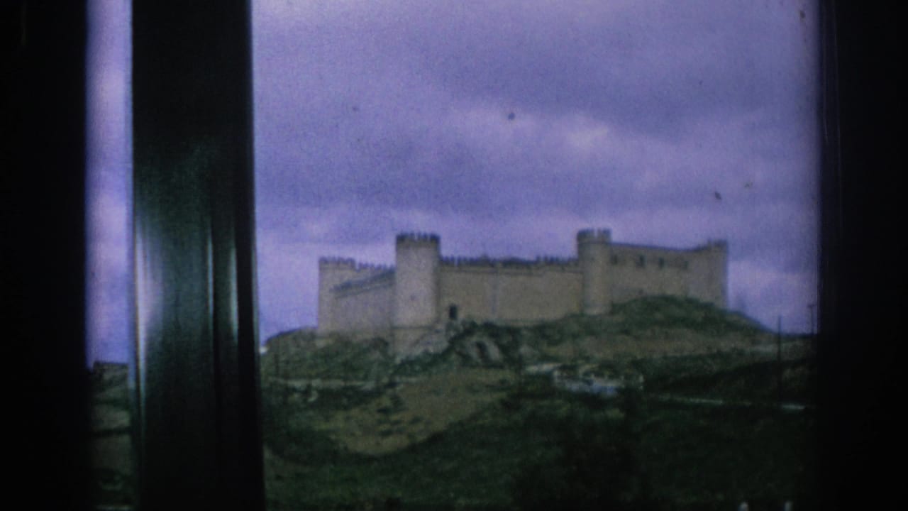 Stone castle on a grassy hill seen through a dark window frame under cloudy sky.
