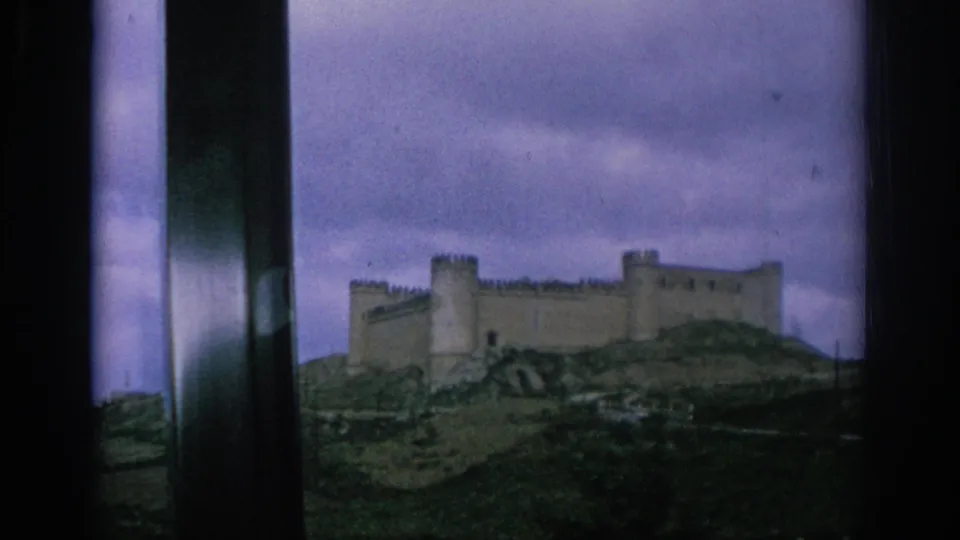 1966 Spain home-movie frame showing the castle’s walls and towers from a moving bus under muted cloudy light.