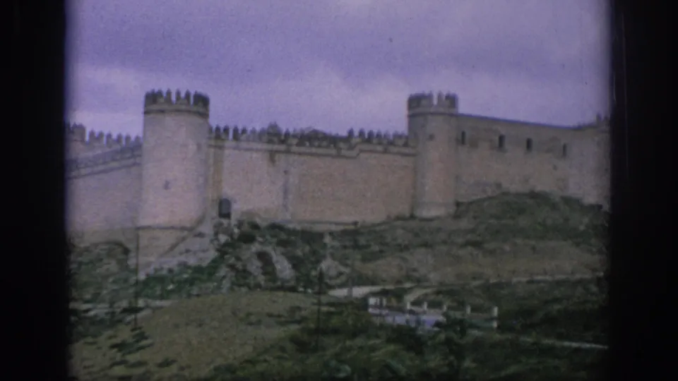 1966 Spain 8mm frame with soft color showing a medieval castle wall, towers, hillside rocks, and sparse vegetation.