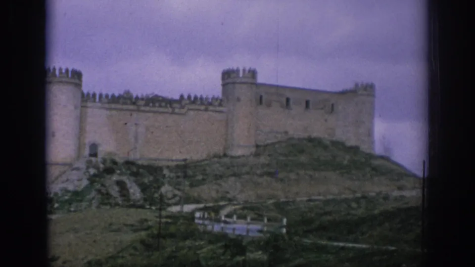 1966 Spain 8mm frame showing a distant hilltop stone castle with crenellated walls, round towers, and dark film borders.