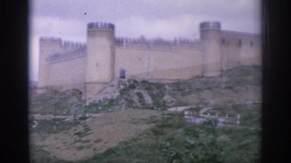 1966 Spain home-movie frame of a pale stone castle wall, round towers, rocky slope, and small path below the fortification.