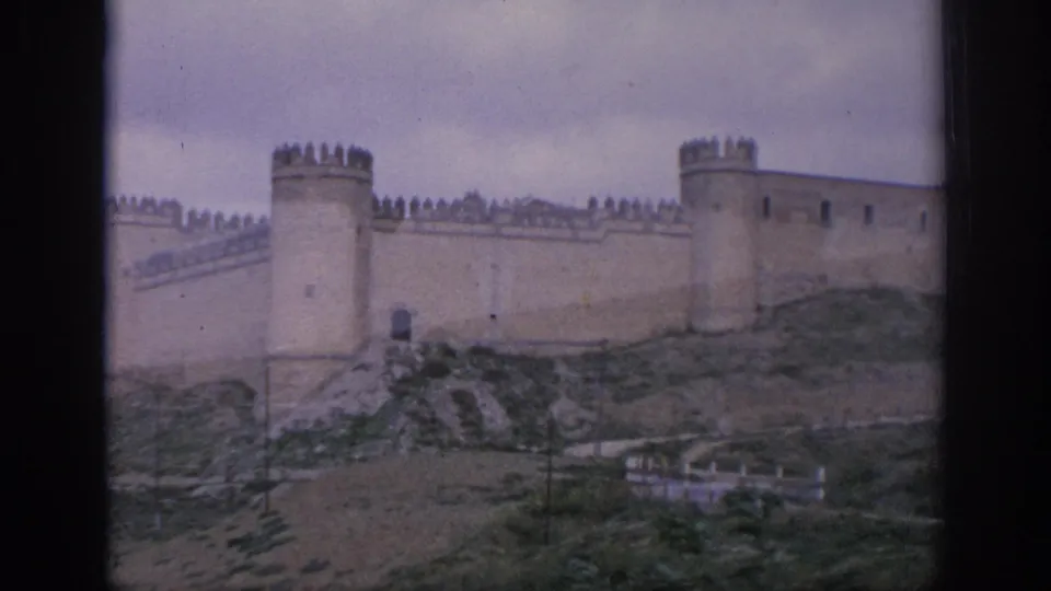 1966 Spain 8mm frame showing crenellated castle walls, a prominent tower, hillside steps or paths, and muted overcast light.