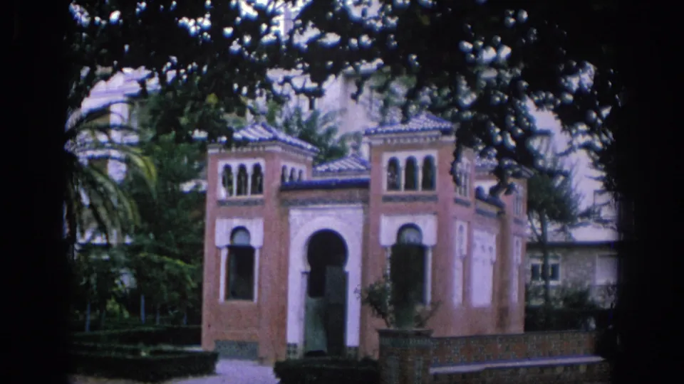 1966 Spain frame of a small garden building with arched openings, patterned roofline, foliage, and dark film edges.