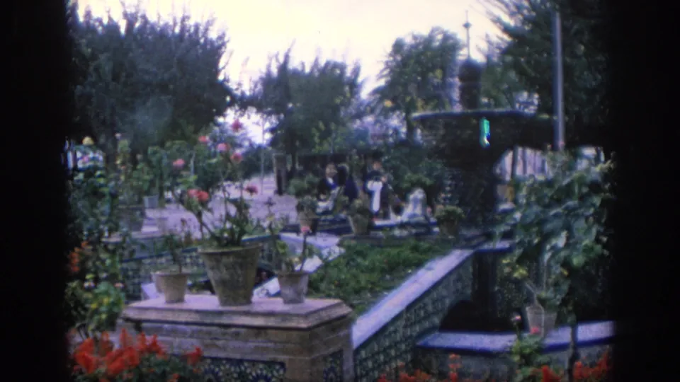 1966 Spain home-movie view of a landscaped terrace with a dark fountain, potted plants, flower beds, tiled edging, and distant visitors.