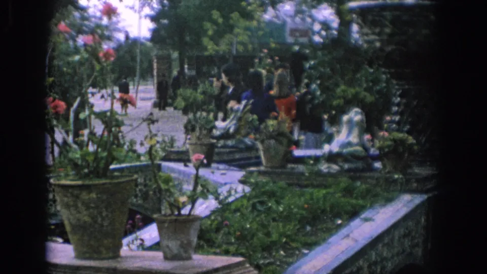 1966 Spain garden footage showing visitors near benches and a fountain, with potted plants, red flowers, tiled walls, and dense greenery.
