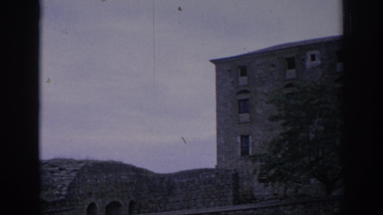 1966 Spain home-movie frame showing a multi-story stone building beside a low wall, with a tree in front and cloudy sky above.