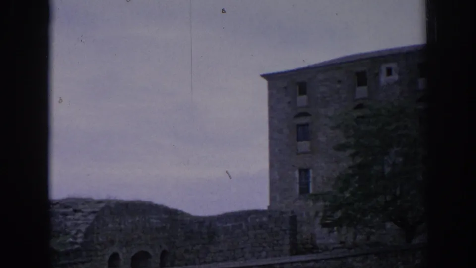 1966 Spain home-movie frame showing a multi-story stone building beside a low wall, with a tree in front and cloudy sky above.