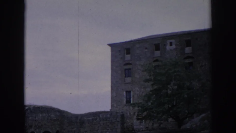 End frame from the 1966 Spain 8mm clip showing the stone parador facade, partial windows, a tree, and low surrounding wall.