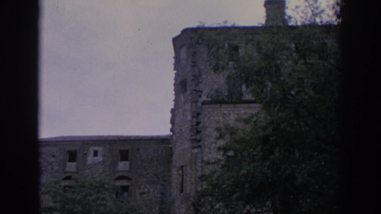1966 Spain 8mm frame showing a weathered stone structure with a rounded corner, dark window openings, and trees in the foreground.