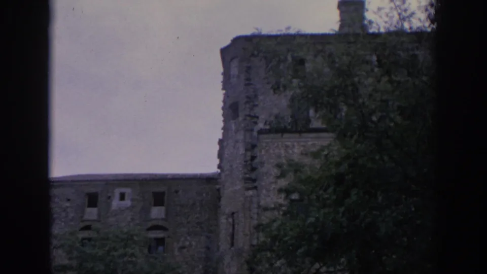 1966 Spain 8mm frame showing a weathered stone structure with a rounded corner, dark window openings, and trees in the foreground.