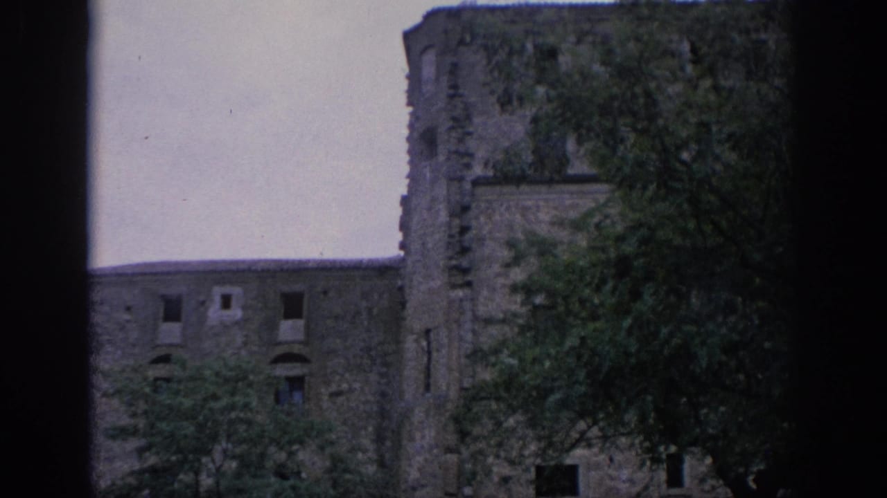 1966 Spain 8mm frame showing an old stone facade with a rounded section, broken-looking openings, and trees partially blocking the view.