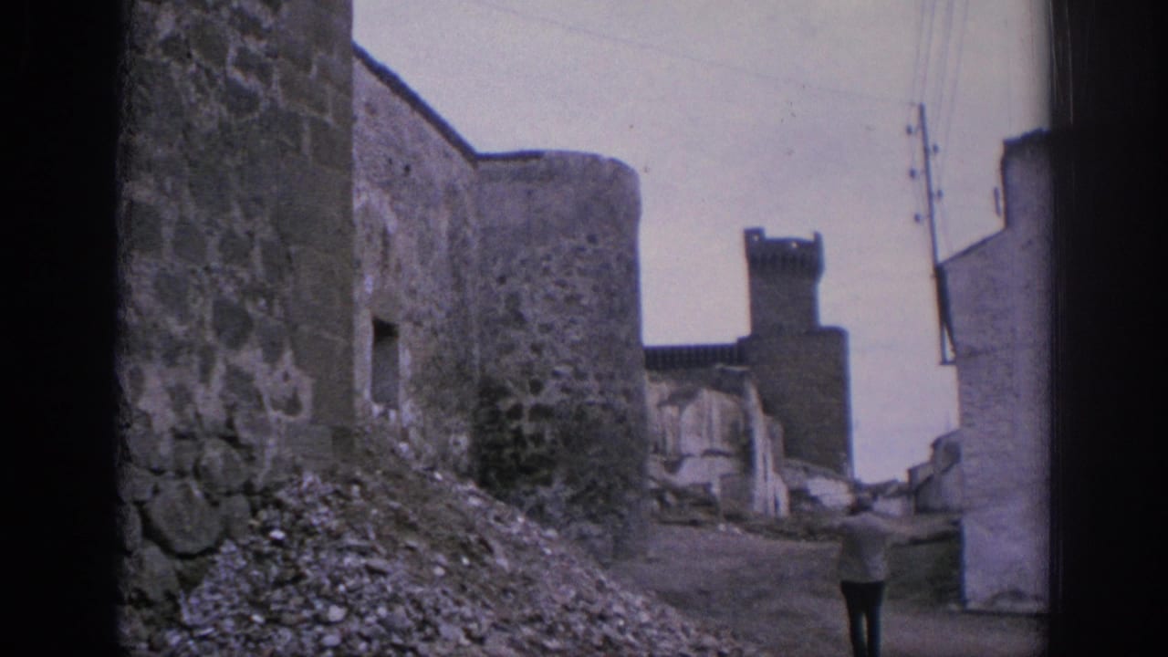1966 Spain 8mm view down a narrow stone-walled lane toward a tower, with rubble, wires, pale buildings, and a distant walker.