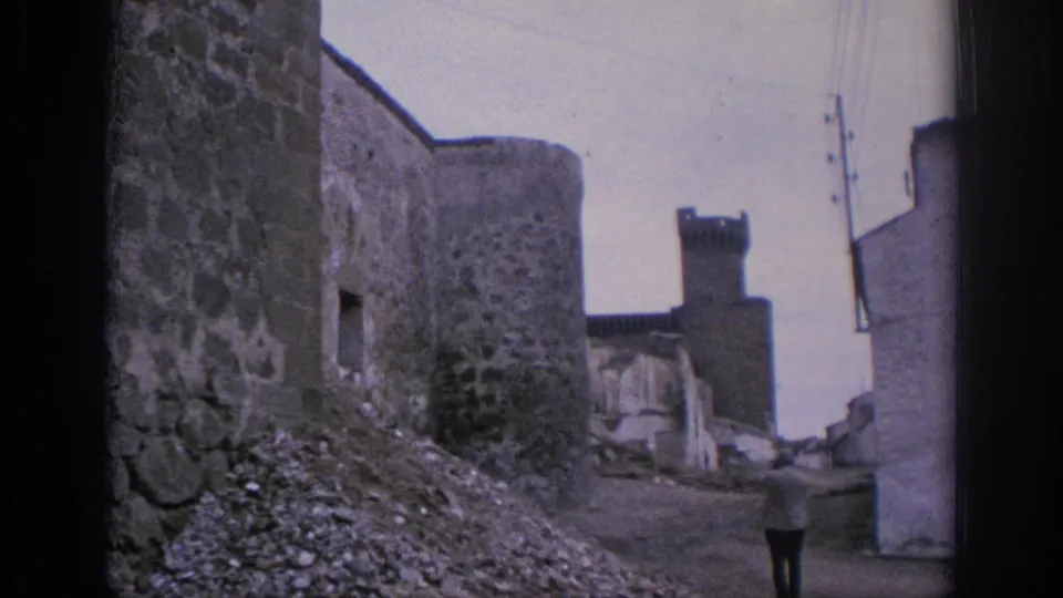 1966 Spain 8mm view down a narrow stone-walled lane toward a tower, with rubble, wires, pale buildings, and a distant walker.