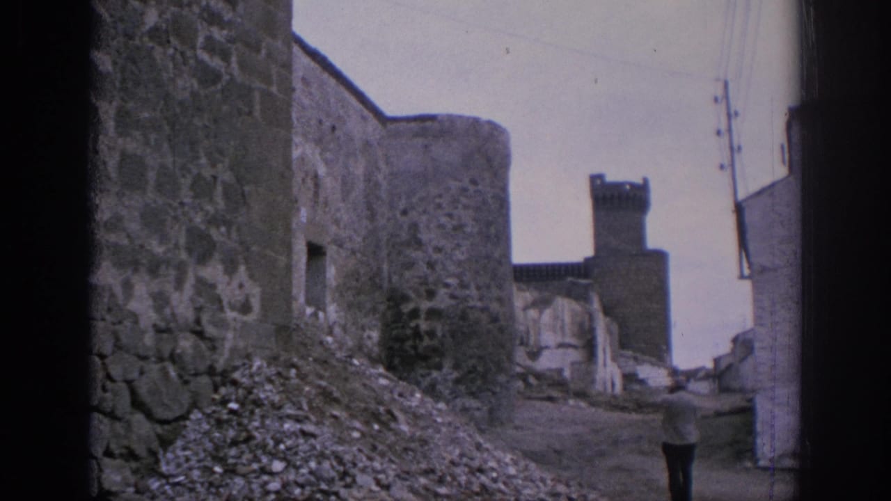 1966 Spain home-movie frame showing a rough stone lane, round tower, gravel debris, nearby buildings, and a distant adult figure.