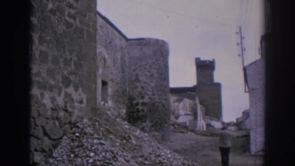 1966 Spain home-movie frame showing a rough stone lane, round tower, gravel debris, nearby buildings, and a distant adult figure.