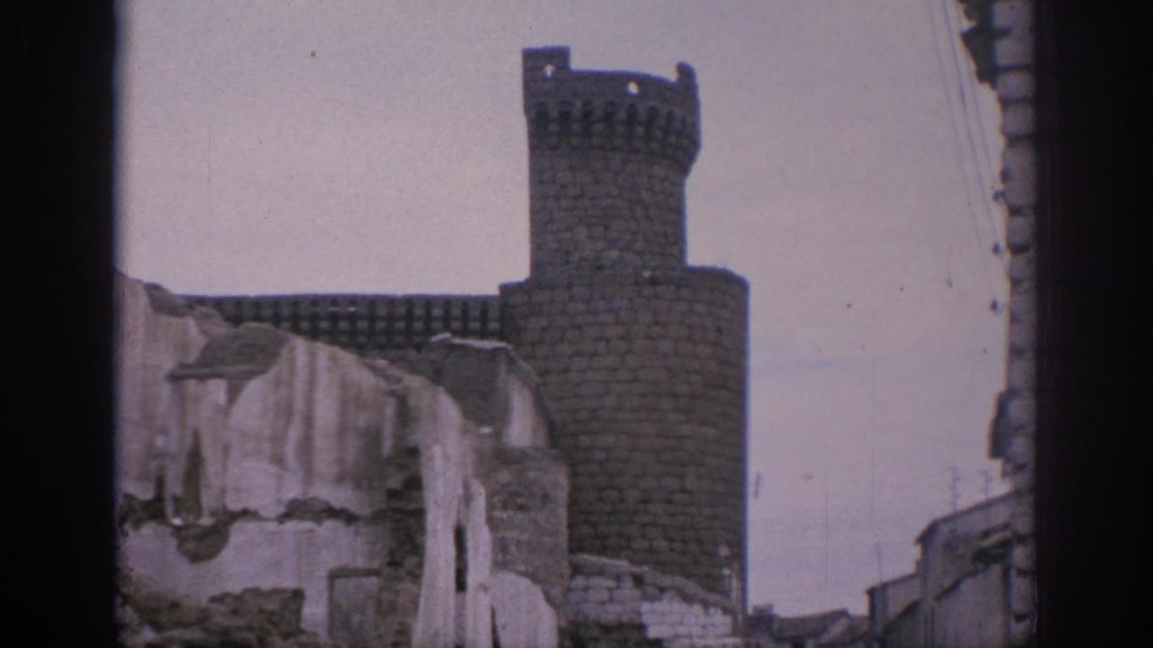 Stone castle tower rising above a damaged pale wall and narrow street edges.