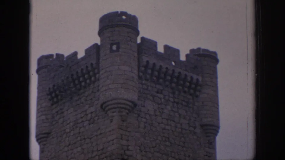 Close 1966 Spain home-movie view of a castle corner with rounded turrets, crenellations, and dark stone masonry.