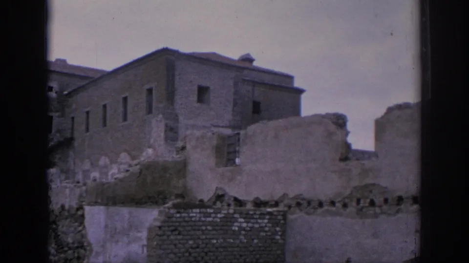 1966 Spain home-movie frame showing a large plain stone building behind broken walls and rough masonry.