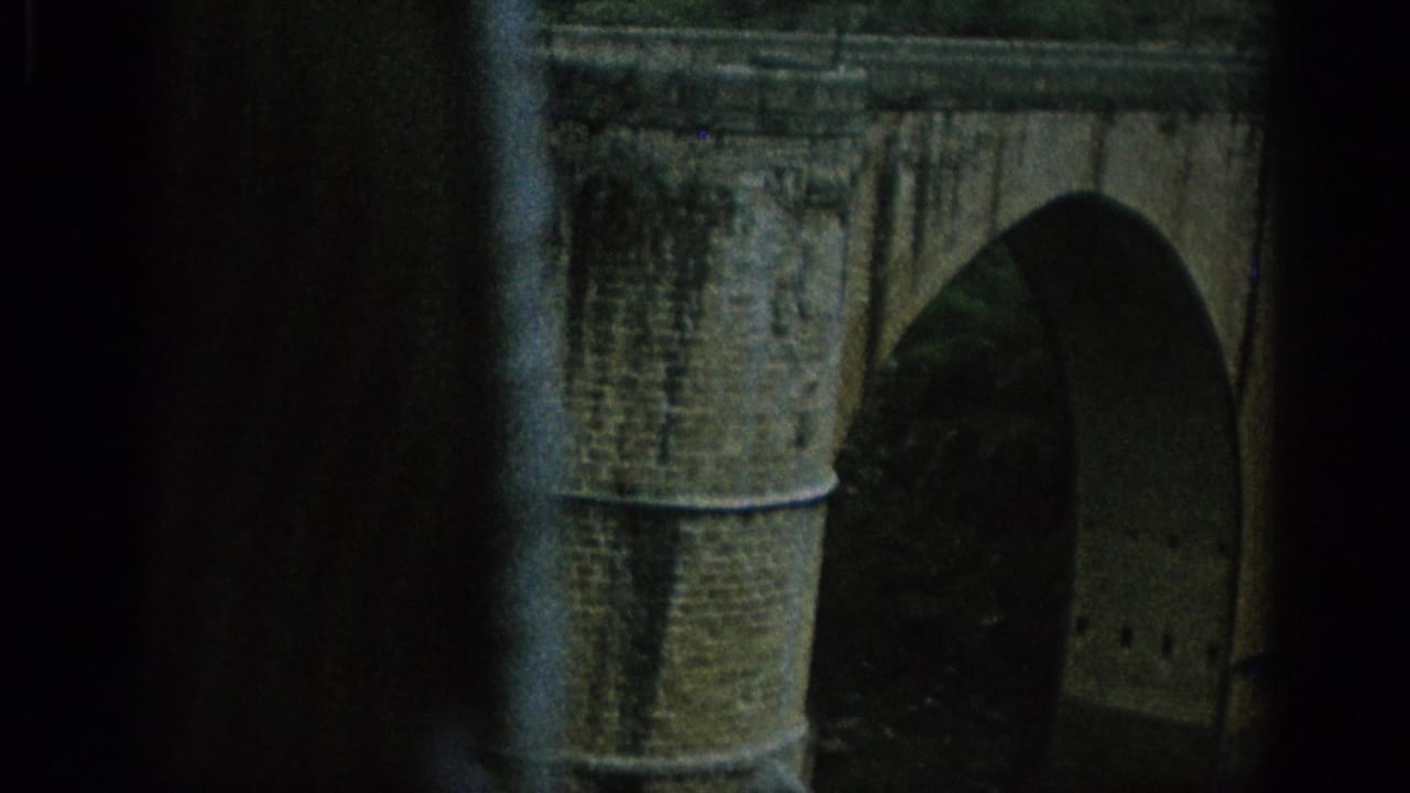 1966 Spain 8mm frame showing a masonry bridge arch in the foreground with a hilltop town and groves beyond.