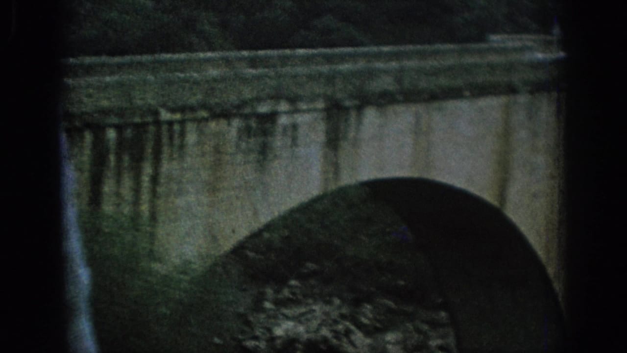 1966 Spain 8mm frame showing a close stone arch bridge, dark window edge, and riverbank vegetation.