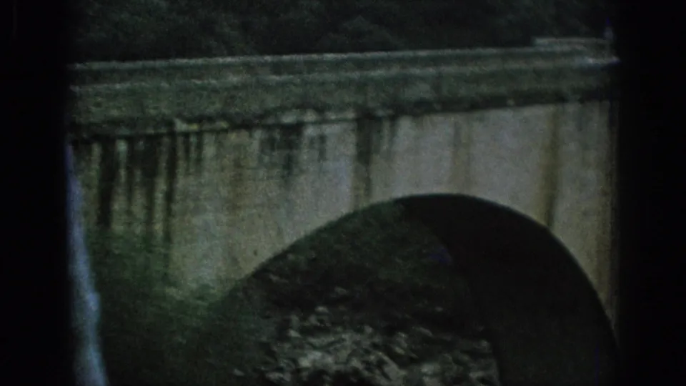 1966 Spain 8mm frame showing a close stone arch bridge, dark window edge, and riverbank vegetation.