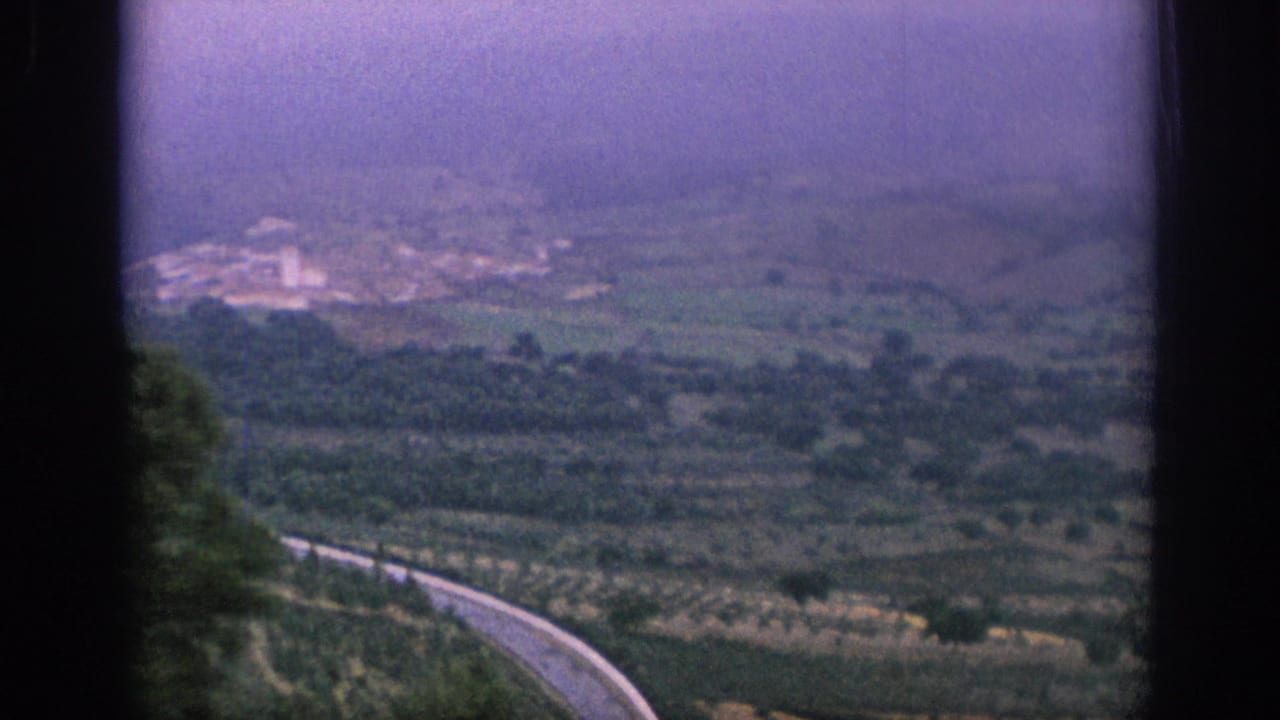 1966 Spain 8mm view from a train over rolling countryside, with a winding road below and hazy hills beyond.
