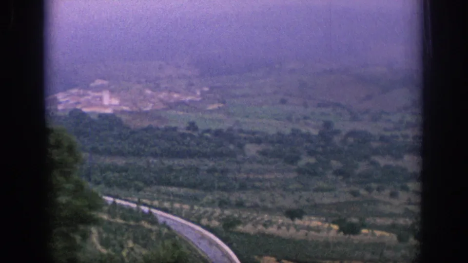 1966 Spain 8mm view from a train over rolling countryside, with a winding road below and hazy hills beyond.