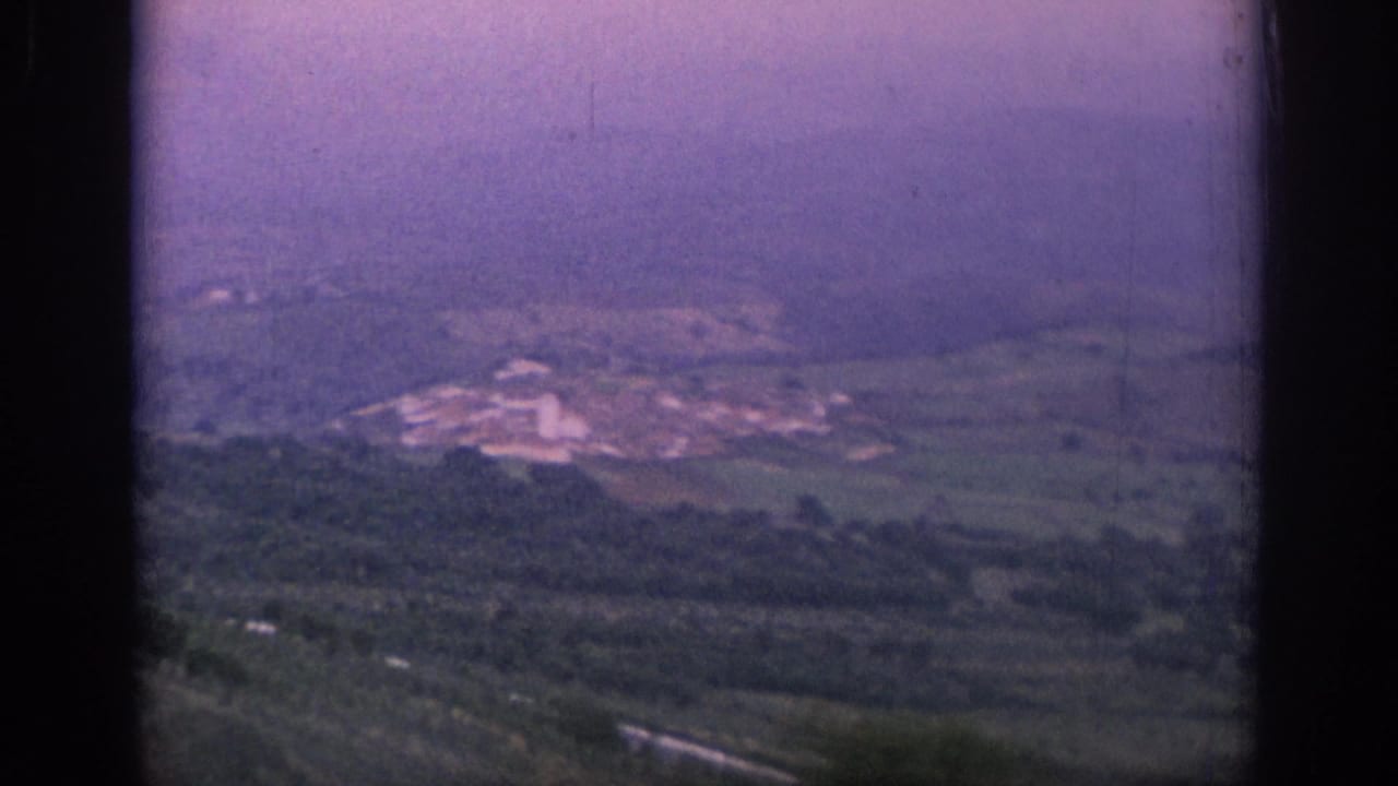 1966 Spain 8mm frame of a hazy countryside panorama, with fields, wooded slopes, and pale buildings in the distance.