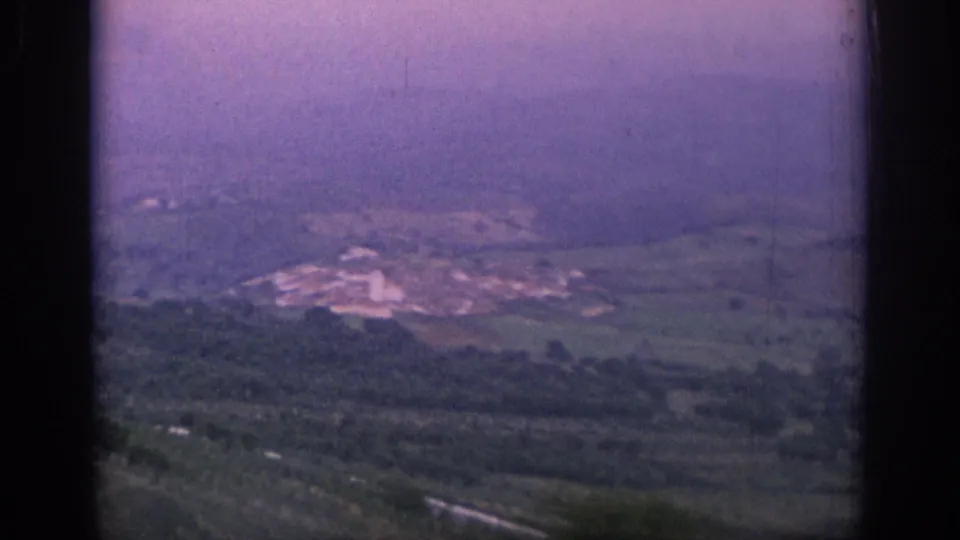 1966 Spain 8mm frame of a hazy countryside panorama, with fields, wooded slopes, and pale buildings in the distance.