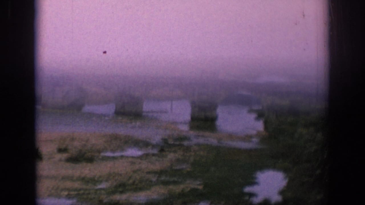 1966 Spain home-movie frame showing a hazy aerial glimpse of a stone arch bridge over a shallow riverbed.