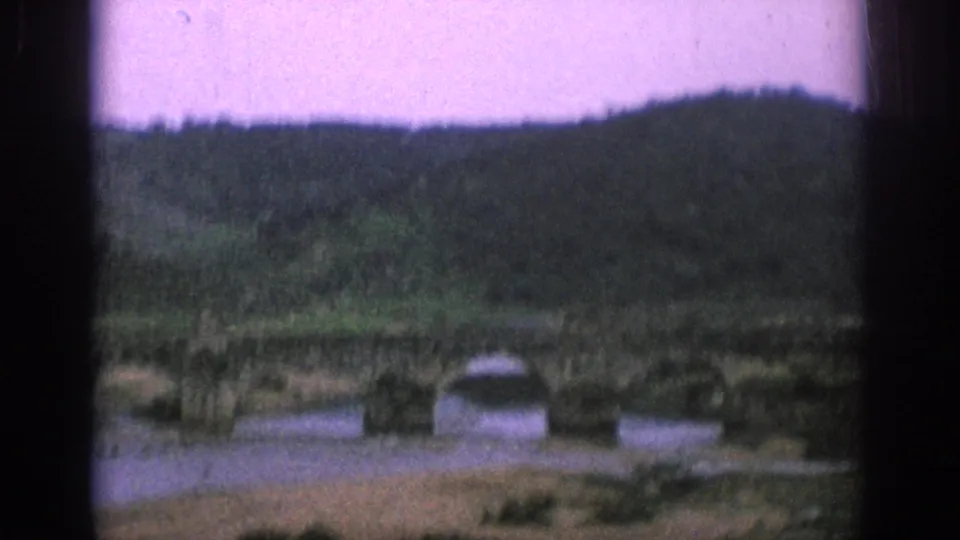 Soft 1966 Spain frame with bridge arches, riverbank, and wooded hill seen from a moving aircraft.