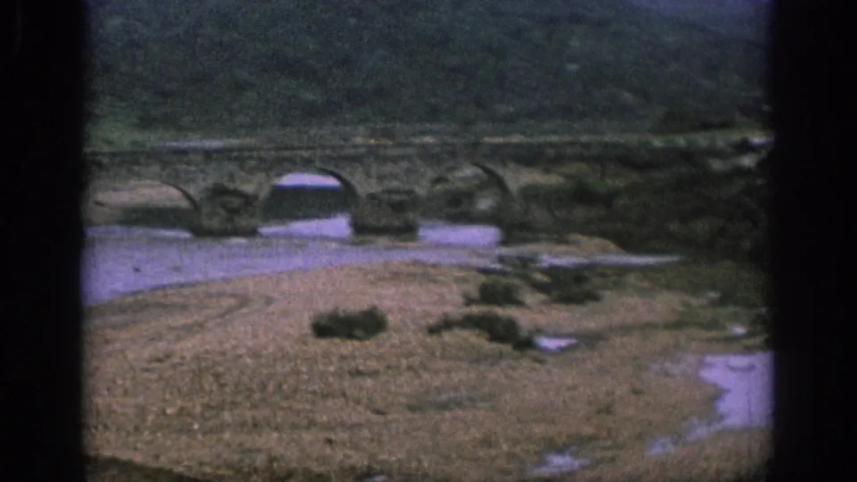 Aerial home-movie frame of a stone arch bridge, shallow water, and gravel bank in a Spanish rural landscape.