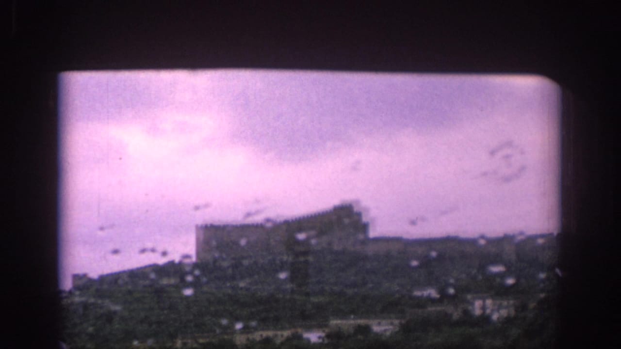 1966 Spain home-movie view through wet train glass toward a distant castle-like structure on a hillside beneath clouds.