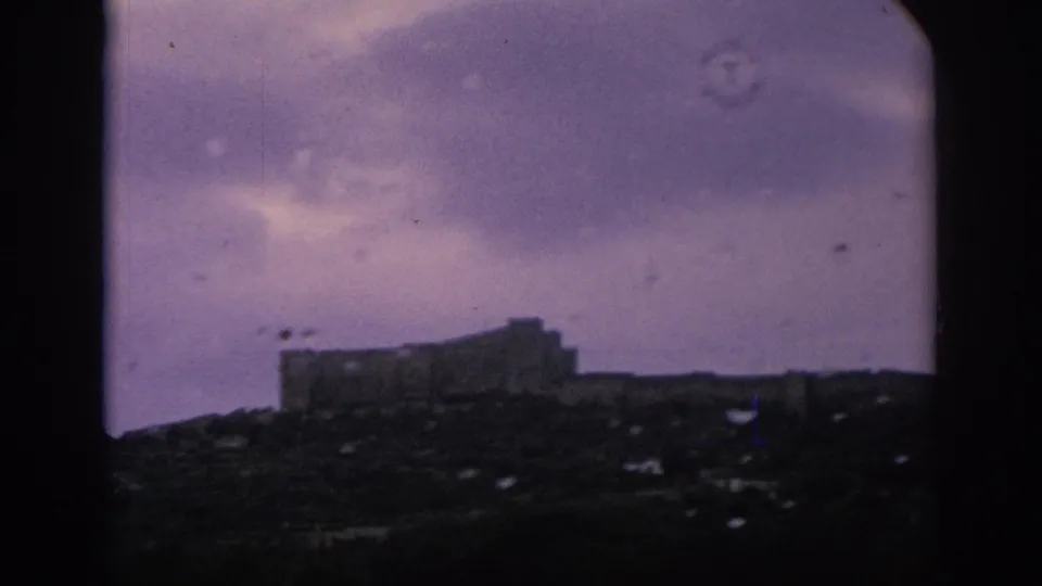 Purple-tinted 8mm frame showing a distant castle-like form on a hillside, viewed through rain-spotted train glass in Spain.