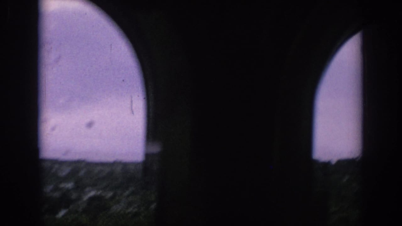 Dark arched window frames a cloudy sky and distant city rooftops through speckled glass.