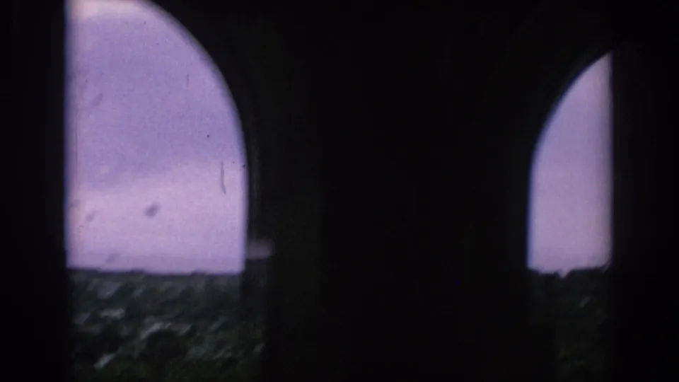1966 Spain home-movie frame looking through dark arched windows toward a cloudy sky and distant urban rooftops.