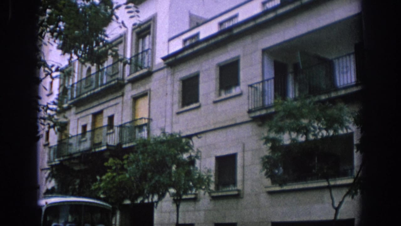 1966 Spain home-movie frame showing a pale residential facade with balconies, trees in front, and part of a white bus below.
