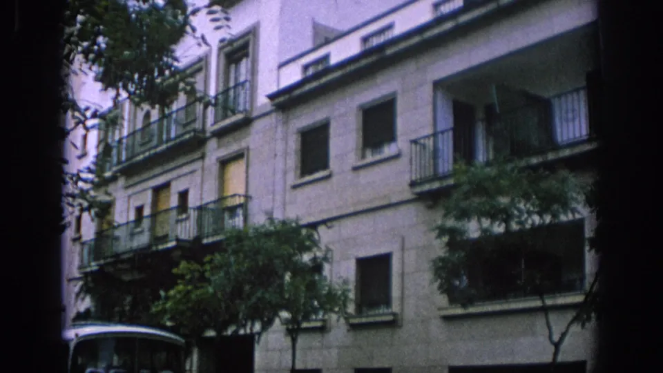 1966 Spain home-movie frame showing a pale residential facade with balconies, trees in front, and part of a white bus below.