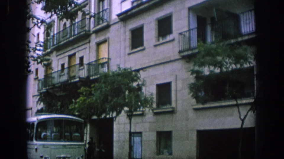 1966 Spain street view with a light-colored apartment block, iron balconies, dark window openings, trees, and a partial bus.