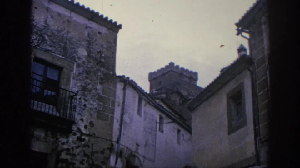 1966 Spain 8mm frame showing weathered stone buildings, barred windows, tiled roofs, and dark film edges in a narrow historic street.