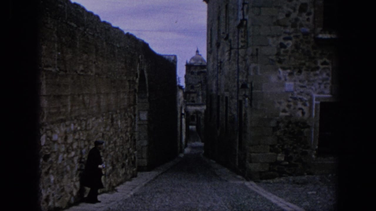 1966 Spain 8mm frame of a narrow cobbled alley, high stone walls, a distant domed structure, and one dark-clothed figure.