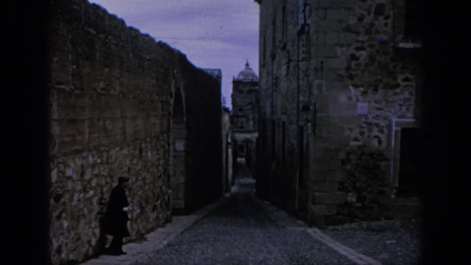 1966 Spain 8mm frame of a narrow cobbled alley, high stone walls, a distant domed structure, and one dark-clothed figure.