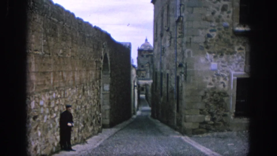 1966 Spain 8mm frame looking down a cobbled alley bordered by old stone walls, with a distant dome and a solitary person near the wall.