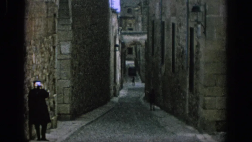 1966 Spain 8mm frame showing a narrow cobbled street between tall stone buildings, with a small distant figure and aged masonry surfaces.