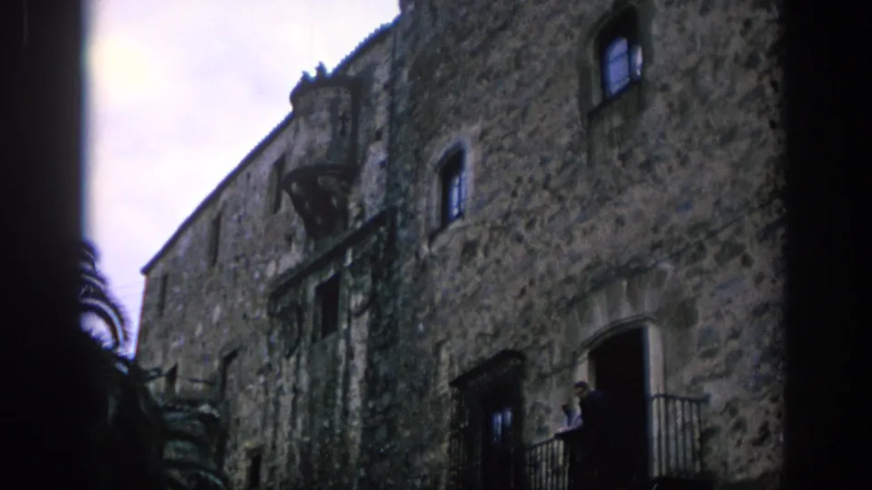 1966 Spain home-movie frame showing a rough stone building facade, small windows, iron balcony, and dark doorway edge.