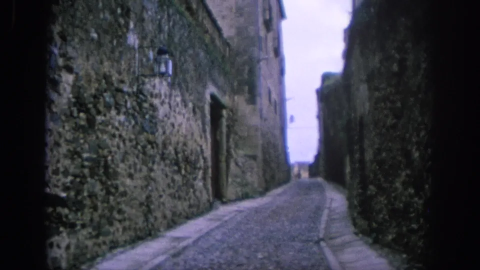 1966 Spain street view down a narrow cobblestone alley bordered by tall stone walls, with a lamp and distant opening visible.